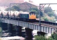 A northbound cement train crosses the doomed bridge over the River Caldew on the Carlisle goods lines shortly after passing through Rome Street Junction in July 1981. The Rome Street gasometer dominates the left background. Damage caused to this bridge by runaway Freightliner vehicles on 1 May 1984 led to final closure of the goods lines. <br><br>[John Furnevel 11/07/1981]