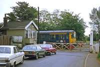 Cars from the 1970s wait on a DMU from the 1950s at Cantley station level crossing on 28th June 1977. The train is a service from Lowestoft to Norwich. Despite major advances in car and train design, the signal box and manual gates endured for decades more to become amongst the last of their kind on the national system.<br><br>[Mark Dufton 28/06/1977]