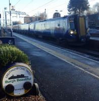 An electric train on the E&G passes a planter at Linlithgow station.<br><br>[John Yellowlees 10/12/2017]